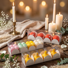 Three clear plastic gift boxes containing assorted decorative candles arranged on a wooden table with lit white taper candles, draped fabric, flowers, and warm blurred lights in the background.