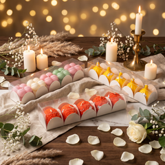 Three clear plastic boxes of decorative bath bombs arranged on a draped beige cloth on a wooden table, surrounded by lit white candles, dried grasses, eucalyptus leaves, rose petals, and a warm golden bokeh background.