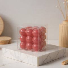 A clear square plastic box containing nine glossy pink spherical items, displayed on a white marble block with neutral wooden props in the background.